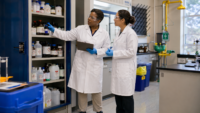 Two scientists in white lab coats and safety goggles examine chemical bottles on a tall shelving unit in a laboratory with hazard labels visible on the bottles