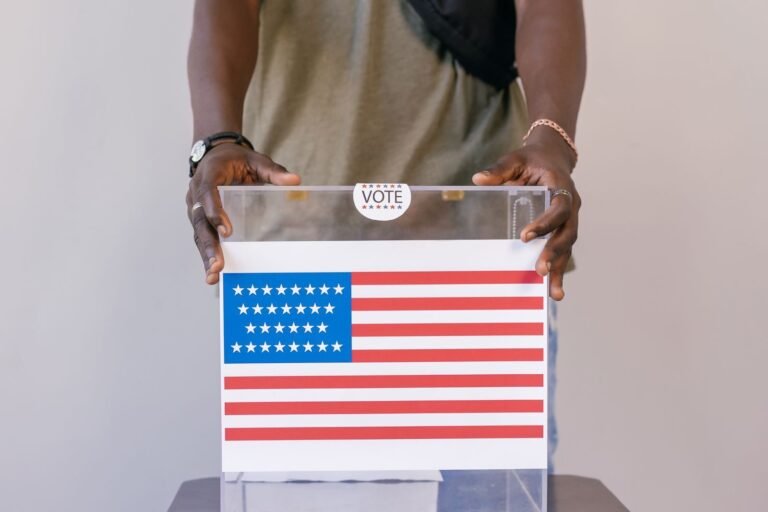 Close-up of a person placing a vote in a transparent ballot box with an American flag prin