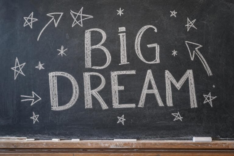 A black chalkboard with the hand-lettered chalk words BIG DREAM surrounded by small doodled stars and arrows, chalk dust on the ledge, soft classroom light