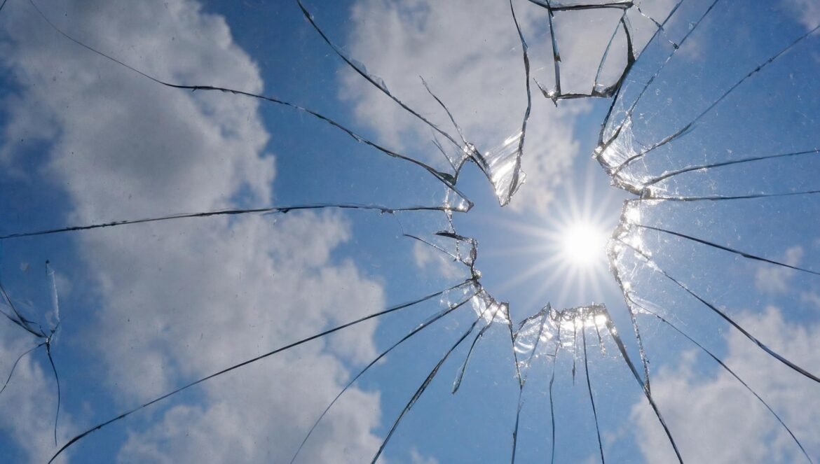 A blue sky with white clouds through a cracked pane of glass bright sun shining through a star shaped hole in the glass