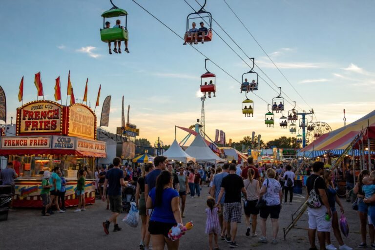 A busy summer state fairground at dusk, families walking along a midway lane under a colorful chairlift sky ride, food stands and tents, warm evening light