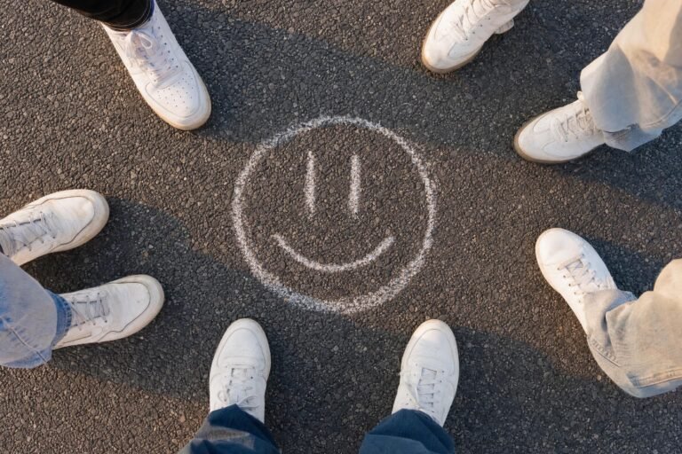 A chalk smiley face drawn on dark asphalt, surrounded by the white sneakers of several people standing in a circle, warm late afternoon light