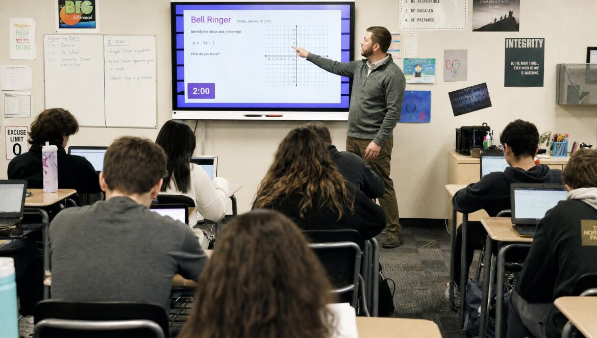 A classroom where students sit at desks working on laptops while a teacher points at a large interactive display screen at the front of the room walls covered