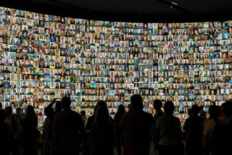 A crowd of people in dark silhouette standing before a huge curved wall covered with hundreds of small glowing portrait photos of faces