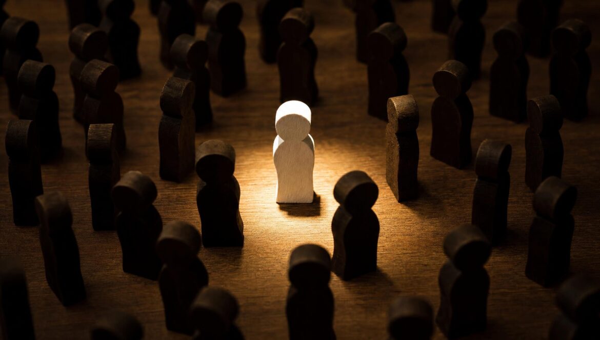 A crowd of small dark wooden figurines on a table with one white figurine in the center lit by a bright spotlight dim warm surroundings | edCircuit A crowd of small dark wooden figurines on a table with one white figurine in the center lit by a bright spotlight dim warm surroundings