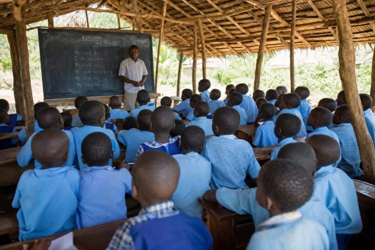 A crowded open-air classroom under a thatched roof, many young children seated on benches facing a teacher, dappled sunlight through wooden poles
