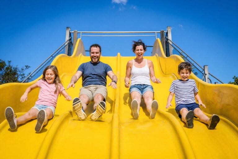 A family of four laughing as they slide down wide bright yellow outdoor slides at a park on a sunny day, blue sky behind