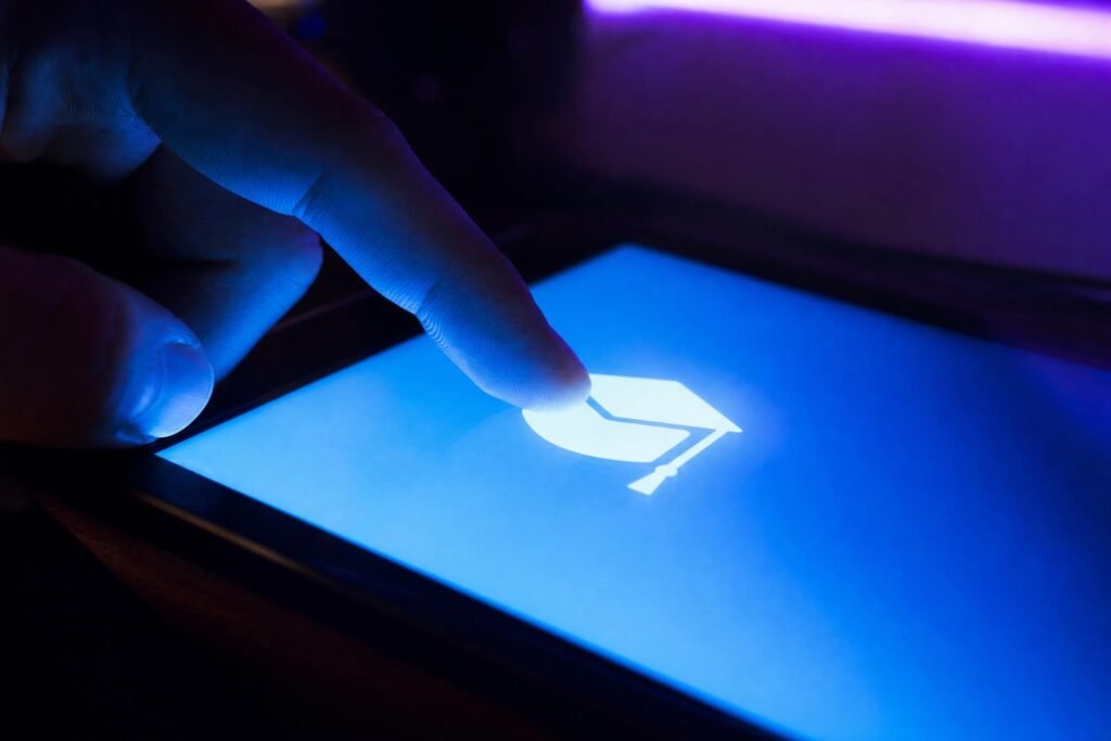 A fingertip touching a glowing blue tablet screen showing a simple graduation cap icon dark room lit by blue and purple screen light | edCircuit A fingertip touching a glowing blue tablet screen showing a simple graduation cap icon dark room lit by blue and purple screen light