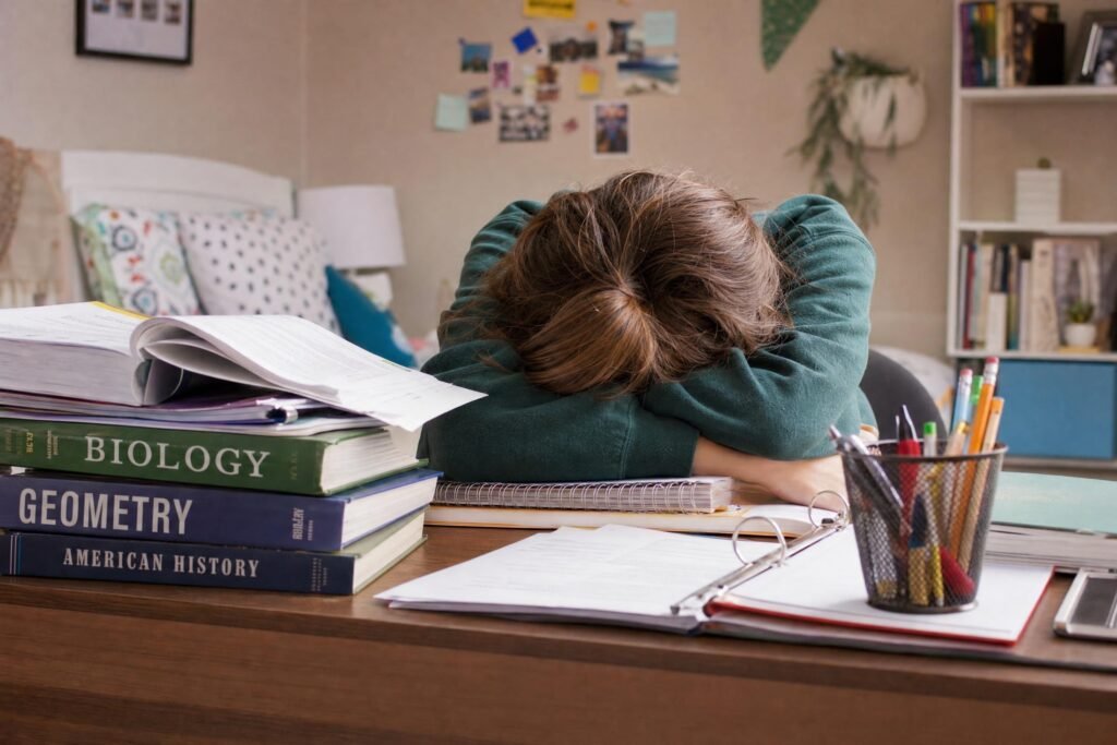 A frustrated student slumped face down on a desk piled with textbooks and a cup of pencils only arms and hair visible colorful dorm room behind | edCircuit A frustrated student slumped face down on a desk piled with textbooks and a cup of pencils only arms and hair visible colorful dorm room behind