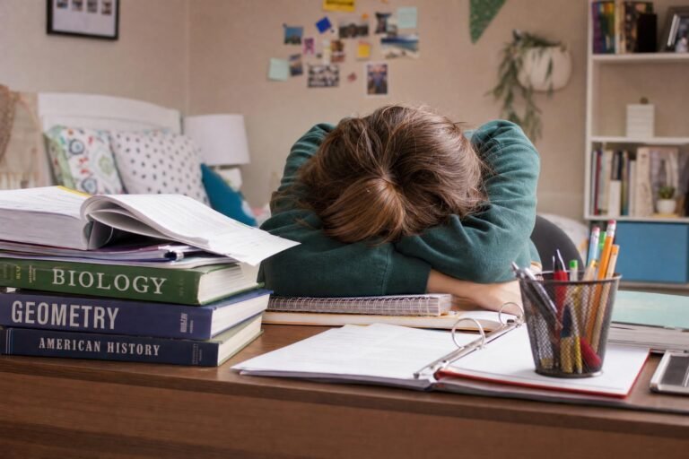 A frustrated student slumped face down on a desk piled with textbooks and a cup of pencils, only arms and hair visible, colorful dorm room behind