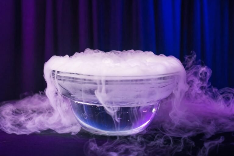 A glass bowl of water with dry ice billowing thick white fog over the rim on a dark table, lit with purple and blue stage light