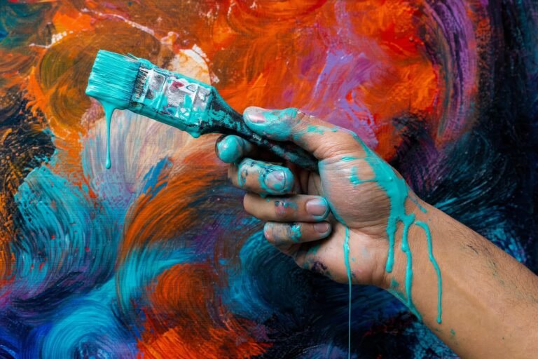 A hand holding a paintbrush with thick turquoise paint dripping down the handle and fingers, in front of a vivid canvas of swirling orange, red, purple and blue