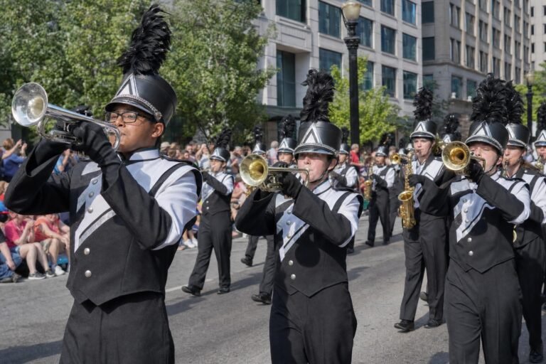 A high school marching band in black and white uniforms playing trumpets while marching down a city street in a parade, spectators behind, bright daylight
