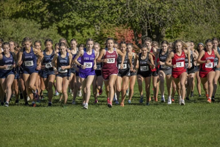 A large pack of teenage girls in team running uniforms starting a cross-country race across a grassy park field, trees behind, bright daylight