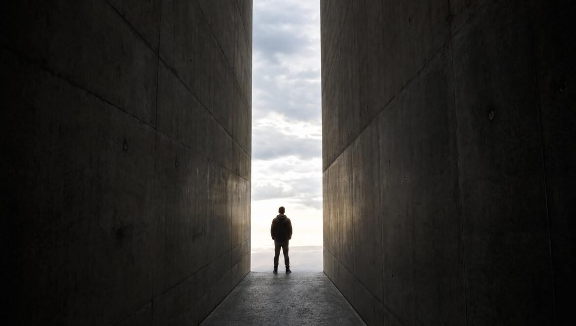A lone person standing in a narrow gap between two towering dark concrete walls bright light and cloudy sky pouring through the opening ahead | edCircuit A lone person standing in a narrow gap between two towering dark concrete walls bright light and cloudy sky pouring through the opening ahead