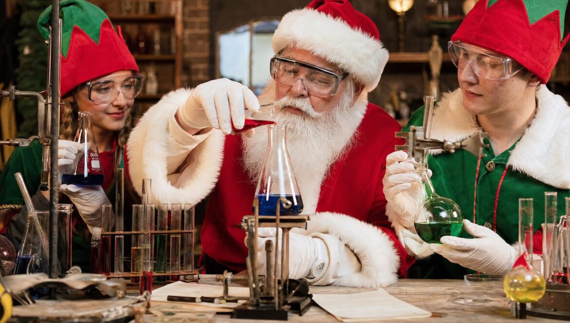 A man dressed as Santa Claus and helpers in elf costumes wearing safety goggles mixing colorful liquids in glass flasks at a cluttered workshop bench warm lam
