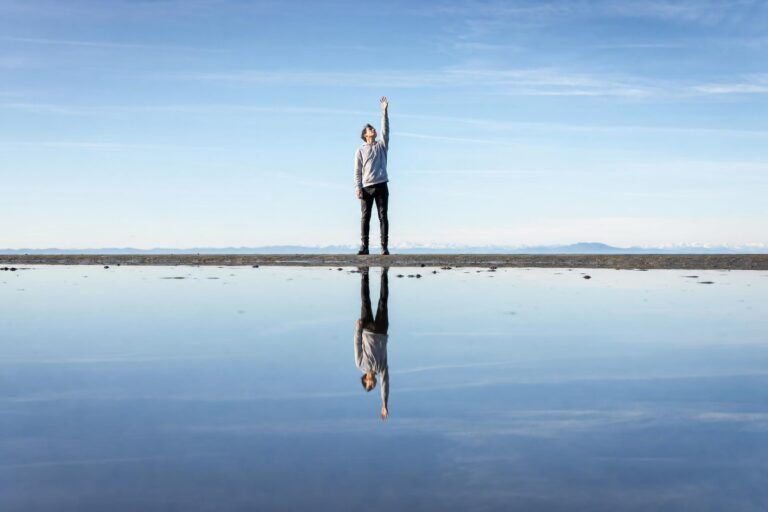 A person standing on a thin wet sandbar reaching one arm up toward a pale blue sky, their figure mirrored in still reflective water below