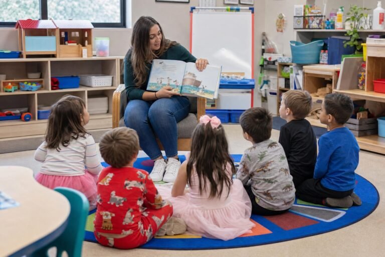 A preschool teacher seated in a chair reading to young children sitting on a colorful round rug in a bright cluttered classroom