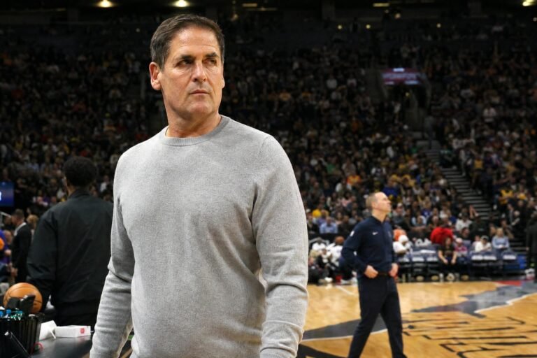 A serious middle-aged man in a gray sweatshirt standing courtside in a busy basketball arena, looking off to the side, warm indoor arena lighting