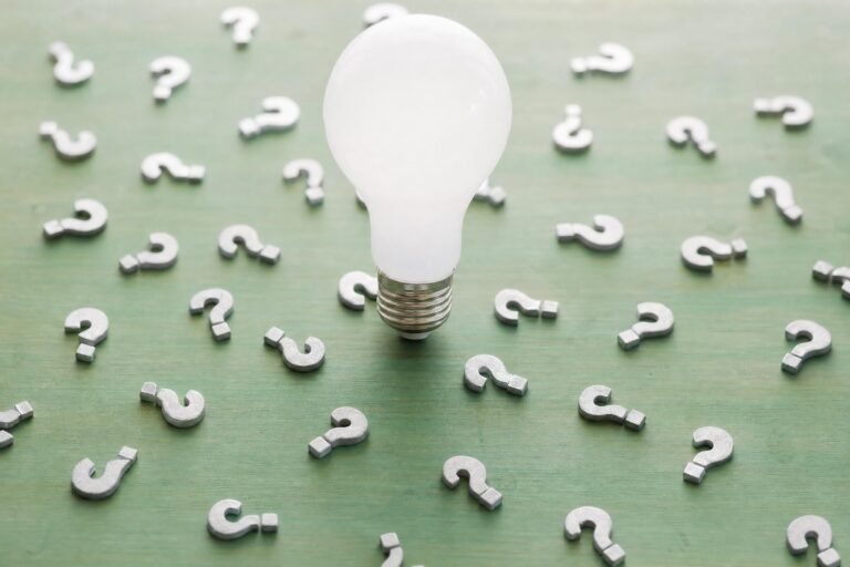 A single white light bulb standing upright on a green table surrounded by scattered small silver question mark shapes, bright soft light
