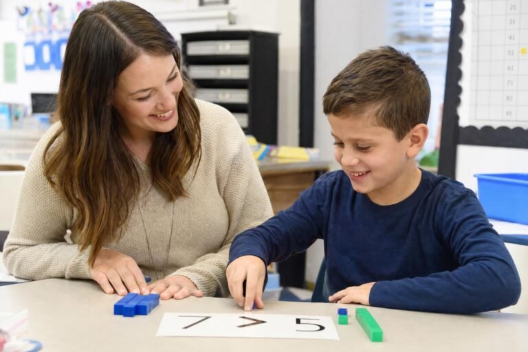 A smiling female teacher sitting at a small table working one on one with a young boy using counting blocks in a warm classroom