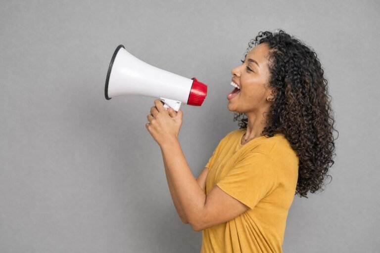 A smiling woman with curly hair shouting into a white megaphone against a plain gray wall, soft studio light