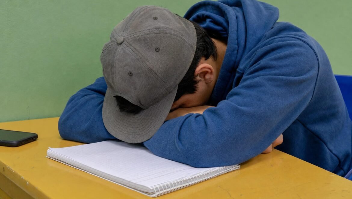 A student in a blue hoodie and gray hat asleep with head down on a yellow desk beside a notebook plain green wall behind | edCircuit A student in a blue hoodie and gray hat asleep with head down on a yellow desk beside a notebook plain green wall behind