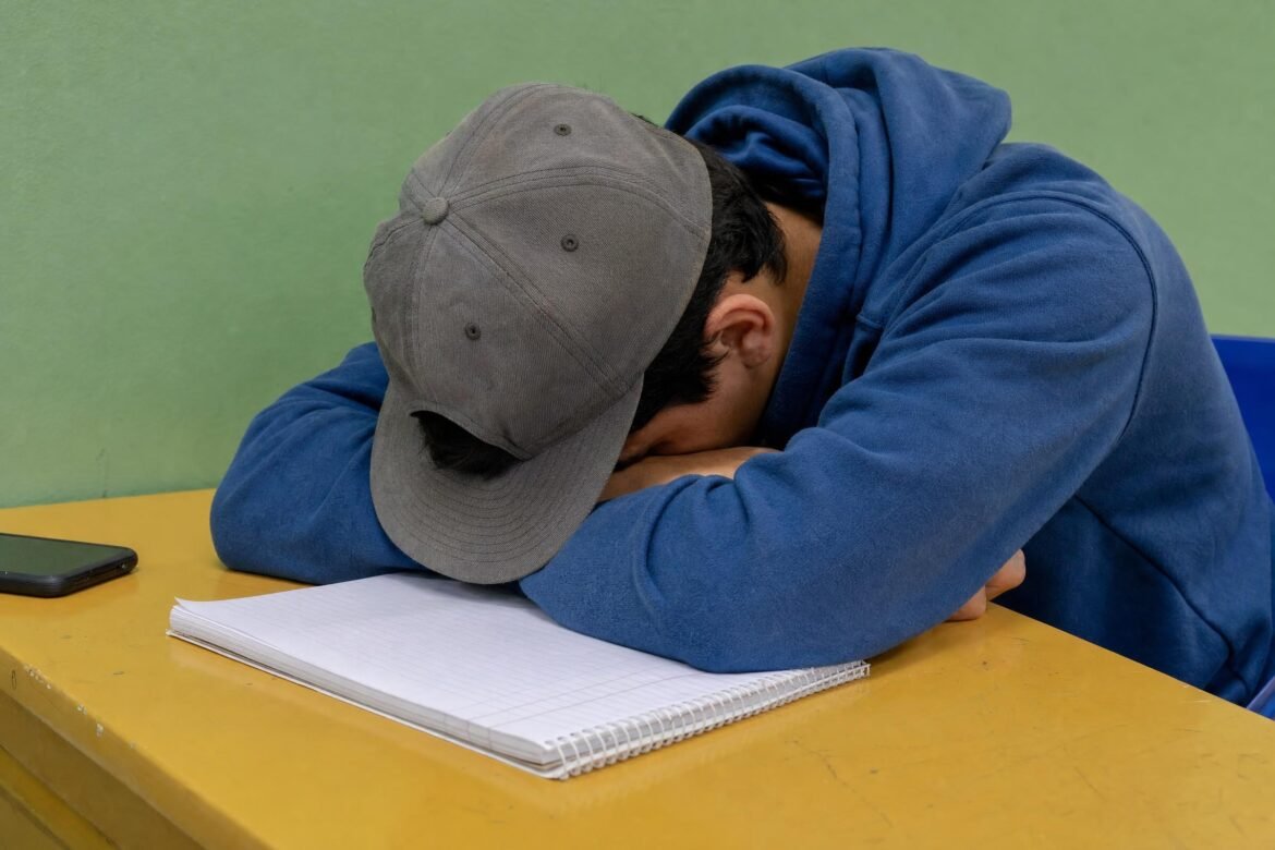 A student in a blue hoodie and gray hat asleep with head down on a yellow desk beside a notebook, plain green wall behind