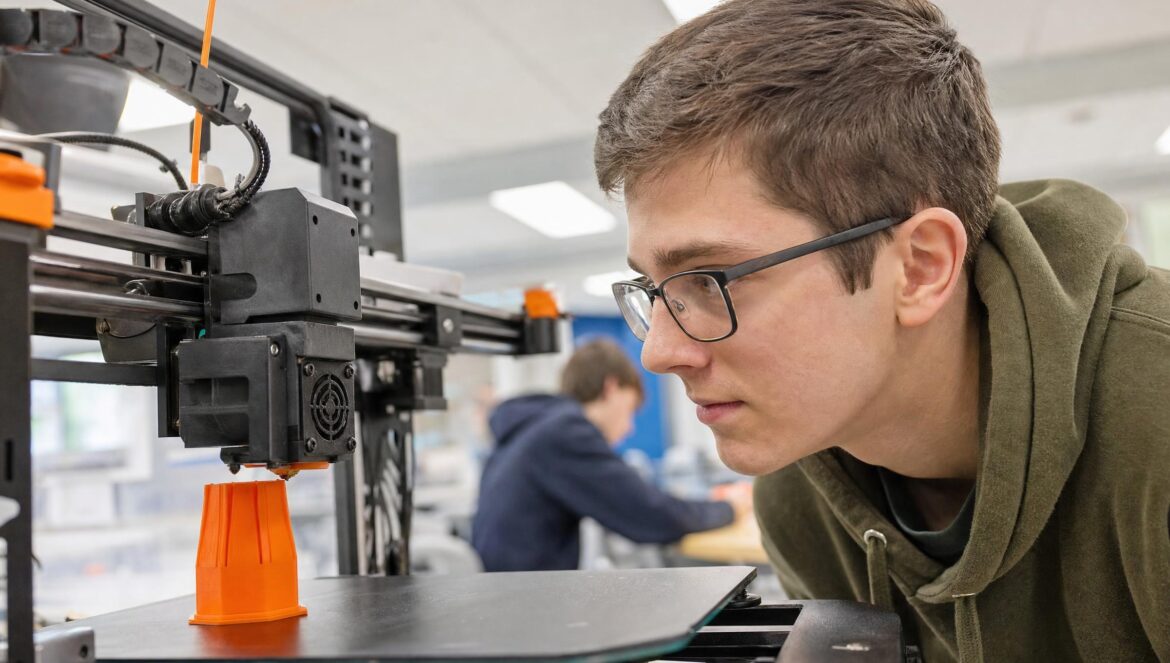 A student in glasses watching closely as a desktop 3D printer builds a small orange plastic part in a bright school makerspace