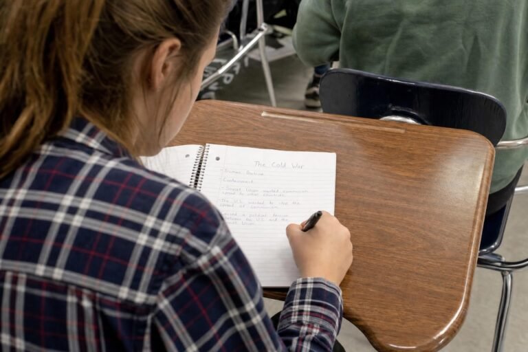 A teenage girl in a checked shirt writing in a lined notebook at her classroom desk, soft natural light