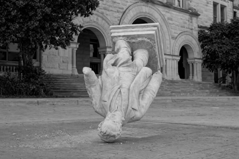 A white marble statue toppled upside down on a stone plaza in front of an arched university building
