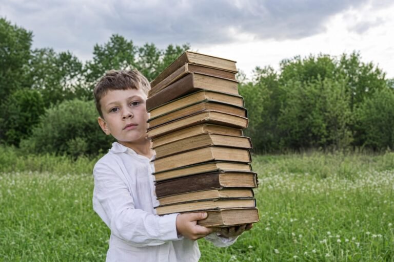 A young boy in a white shirt standing in a green meadow holding a tall wobbly stack of old books in his outstretched arms, trees and cloudy sky behind