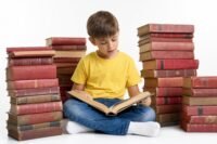 A young boy in a yellow shirt sitting cross-legged with an open book on his lap, surrounded by tall leaning stacks of old red and brown hardcover books, bright