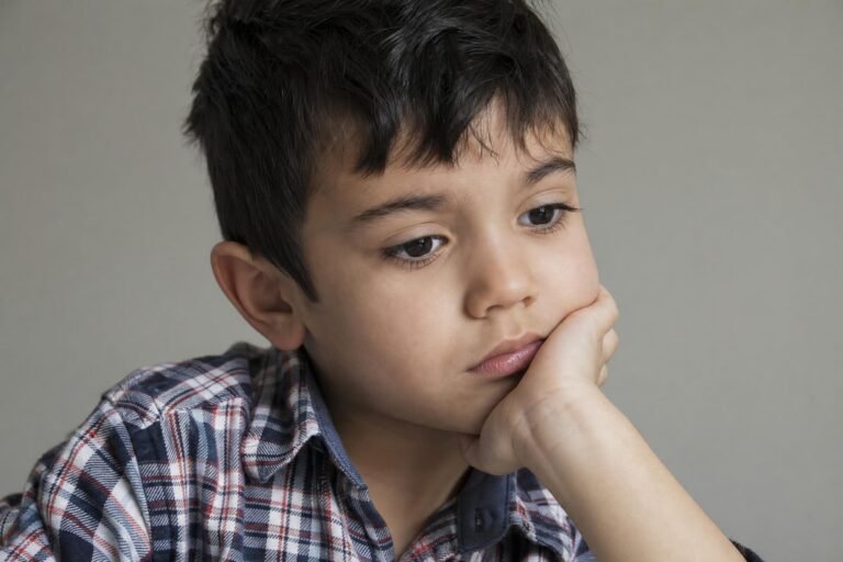 A young dark-haired boy resting his cheek on his hand with a sad thoughtful expression, plaid shirt, plain gray background, soft window light