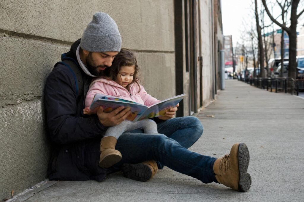 A young father in a knit cap sitting on a city sidewalk against a wall reading a picture book to his small daughter on his lap soft daylight | edCircuit A young father in a knit cap sitting on a city sidewalk against a wall reading a picture book to his small daughter on his lap soft daylight