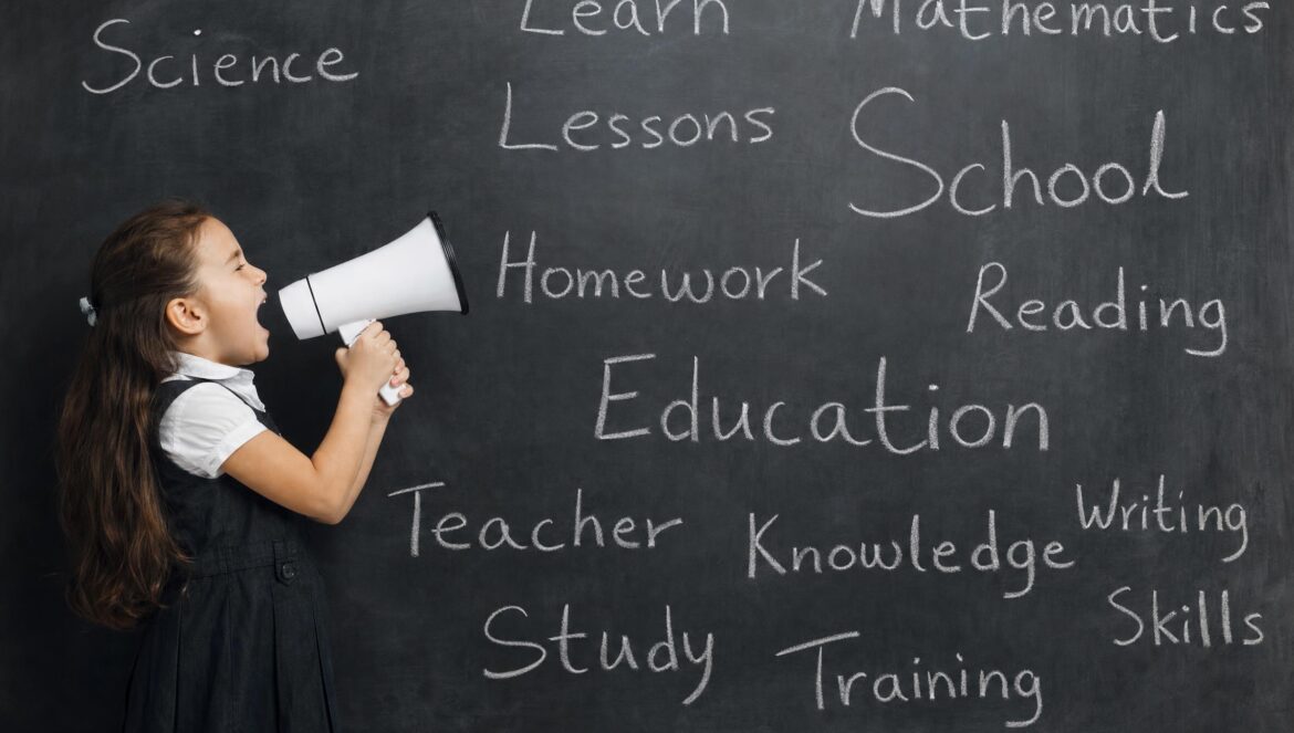 A young girl shouting into a white megaphone beside a large chalkboard covered in chalk words about school and learning even classroom light