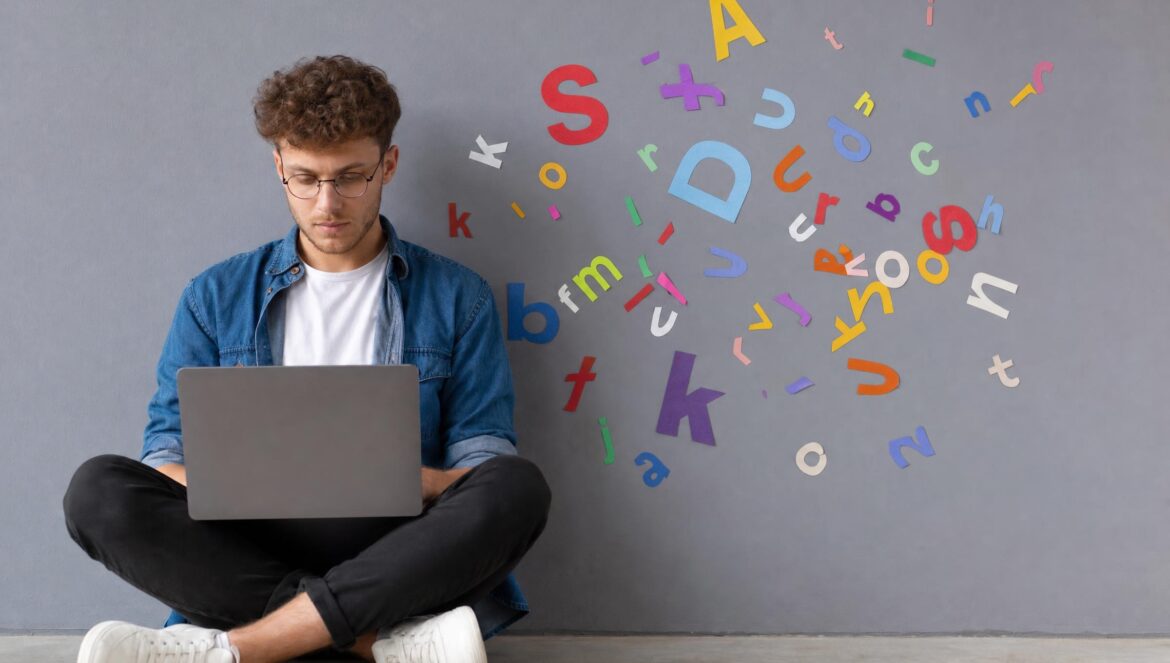 A young man sitting on a concrete floor against a grey wall typing on a laptop colorful paper cutout letters scattered in the air beside him soft even studio | edCircuit A young man sitting on a concrete floor against a grey wall typing on a laptop colorful paper cutout letters scattered in the air beside him soft even studio