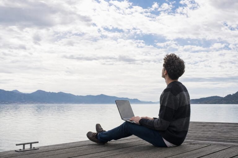 A young man sitting on a wooden pier with a laptop on his knees, gazing up at a wide cloudy sky, calm hazy daylight