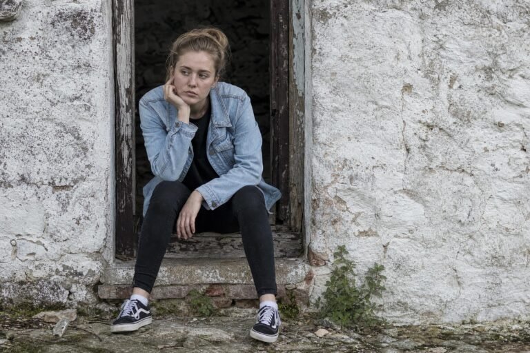 A young woman in a denim jacket sitting hunched in the doorway of an old whitewashed wall, face resting in her hand, soft overcast daylight
