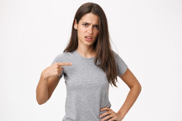 A young woman in a grey t-shirt with a frustrated, questioning expression pointing at herself with one hand on her hip, plain white studio background