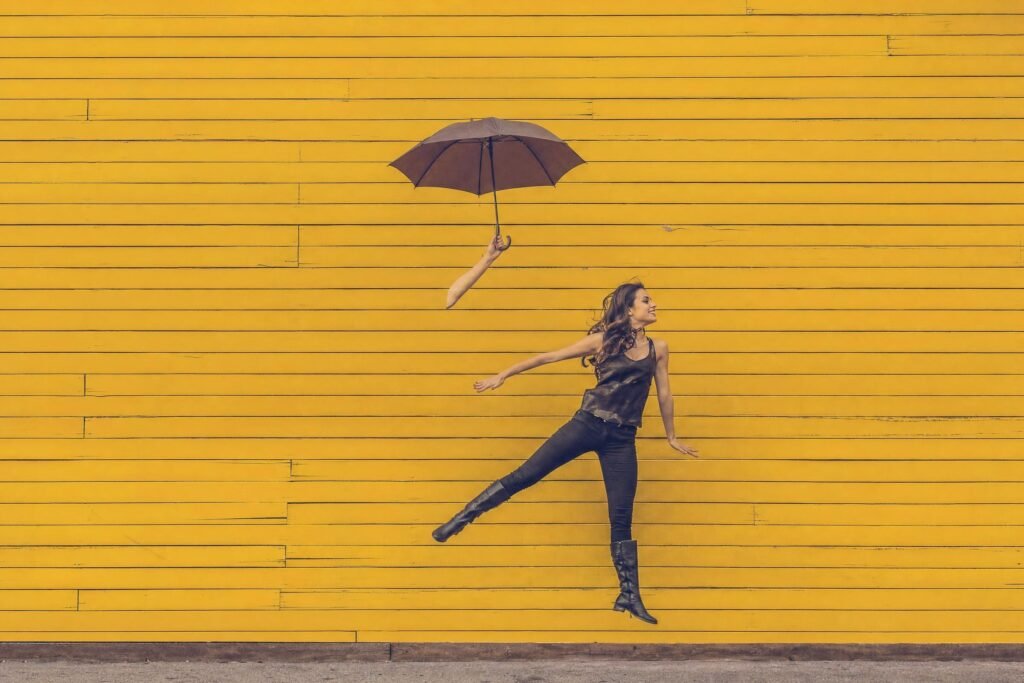 A young woman in dark clothes leaping upward while holding an open umbrella in front of a bright yellow painted wooden wall as if floating in soft daylight | edCircuit A young woman in dark clothes leaping upward while holding an open umbrella in front of a bright yellow painted wooden wall as if floating in soft daylight