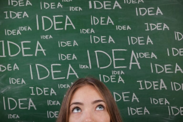 A young woman peeking up from the bottom of the frame in front of a green chalkboard covered with the word IDEA written many times in white chalk, soft classroo