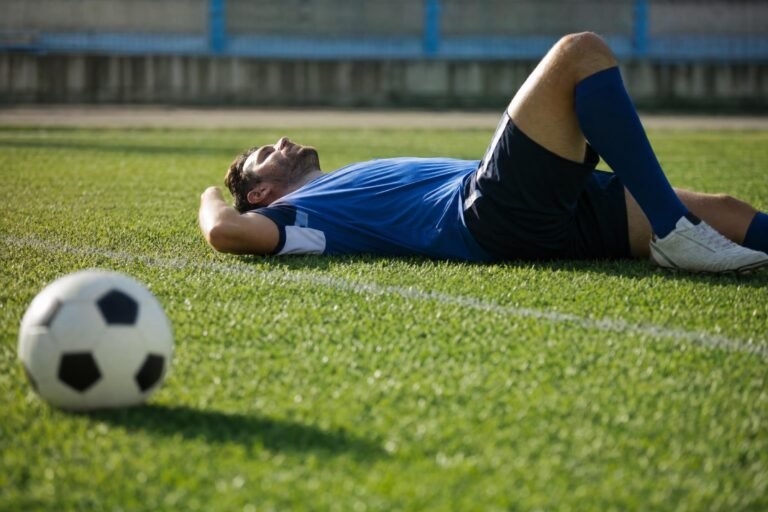 An adult soccer player lying on his back resting on a green grass field, a soccer ball in the foreground, bright afternoon sunlight, shallow depth of field