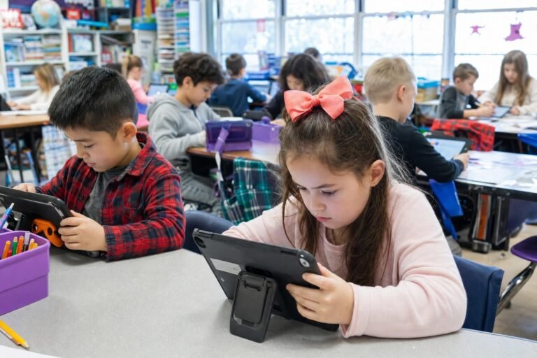 Elementary students working on tablets at shared desks in a busy colorful classroom a girl with a hair bow in front bright daylight | edCircuit Elementary students working on tablets at shared desks in a busy colorful classroom a girl with a hair bow in front bright daylight