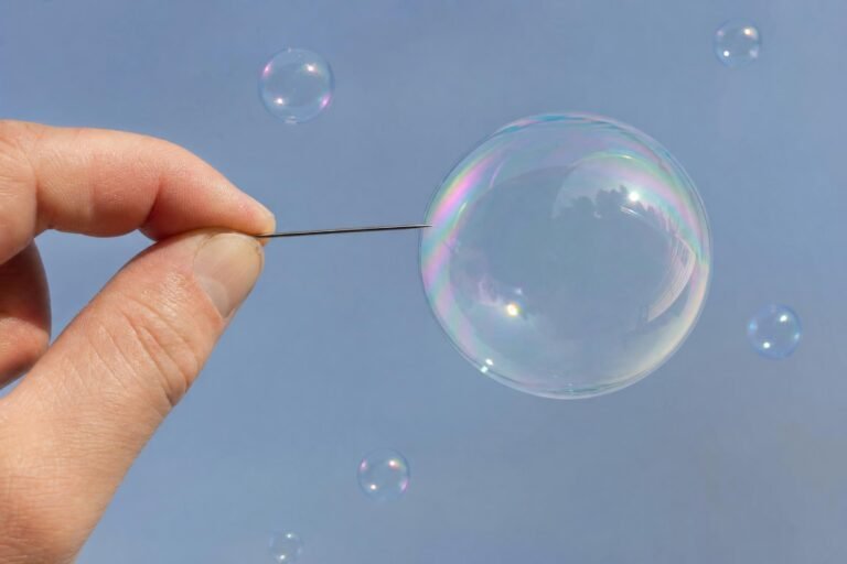 Fingers holding a sewing needle about to pop a large shimmering soap bubble, several smaller bubbles floating nearby against a soft pale blue background