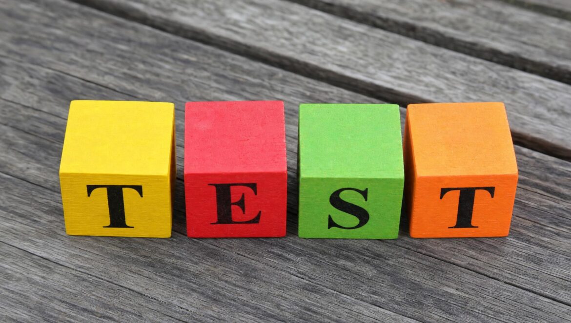 Four colorful wooden toy blocks in yellow red green and orange lined up on a weathered grey wooden table painted black letters spelling TEST soft daylight | edCircuit Four colorful wooden toy blocks in yellow red green and orange lined up on a weathered grey wooden table painted black letters spelling TEST soft daylight