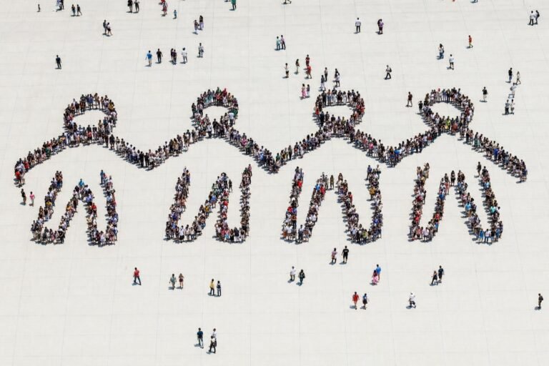 From high above of hundreds of people standing in a white plaza arranged to form giant human figures holding hands, bright overhead daylight