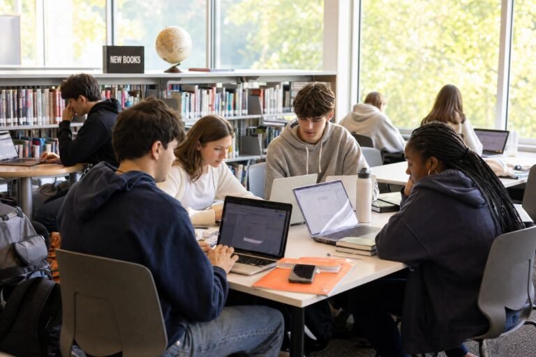 High school students working on laptops around white tables in a bright school library, warm afternoon light