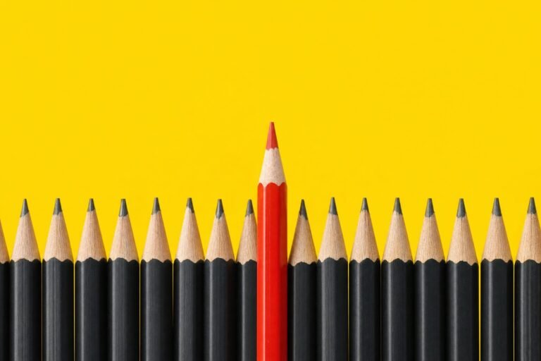 One red pencil standing taller in the middle of a long row of black pencils against a bright yellow background