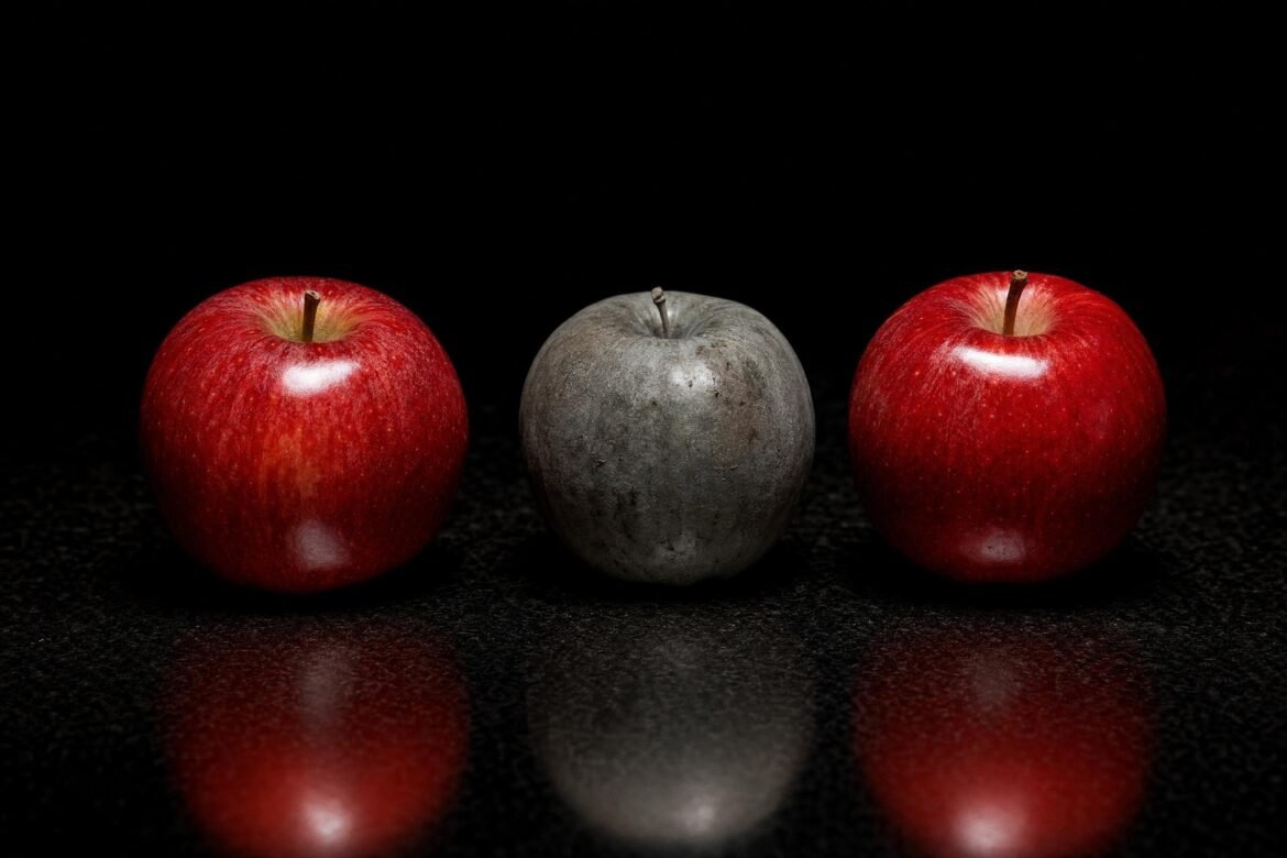 Three apples in a row on a black reflective surface, two shiny red apples flanking one dull gray apple in the middle, dramatic top light